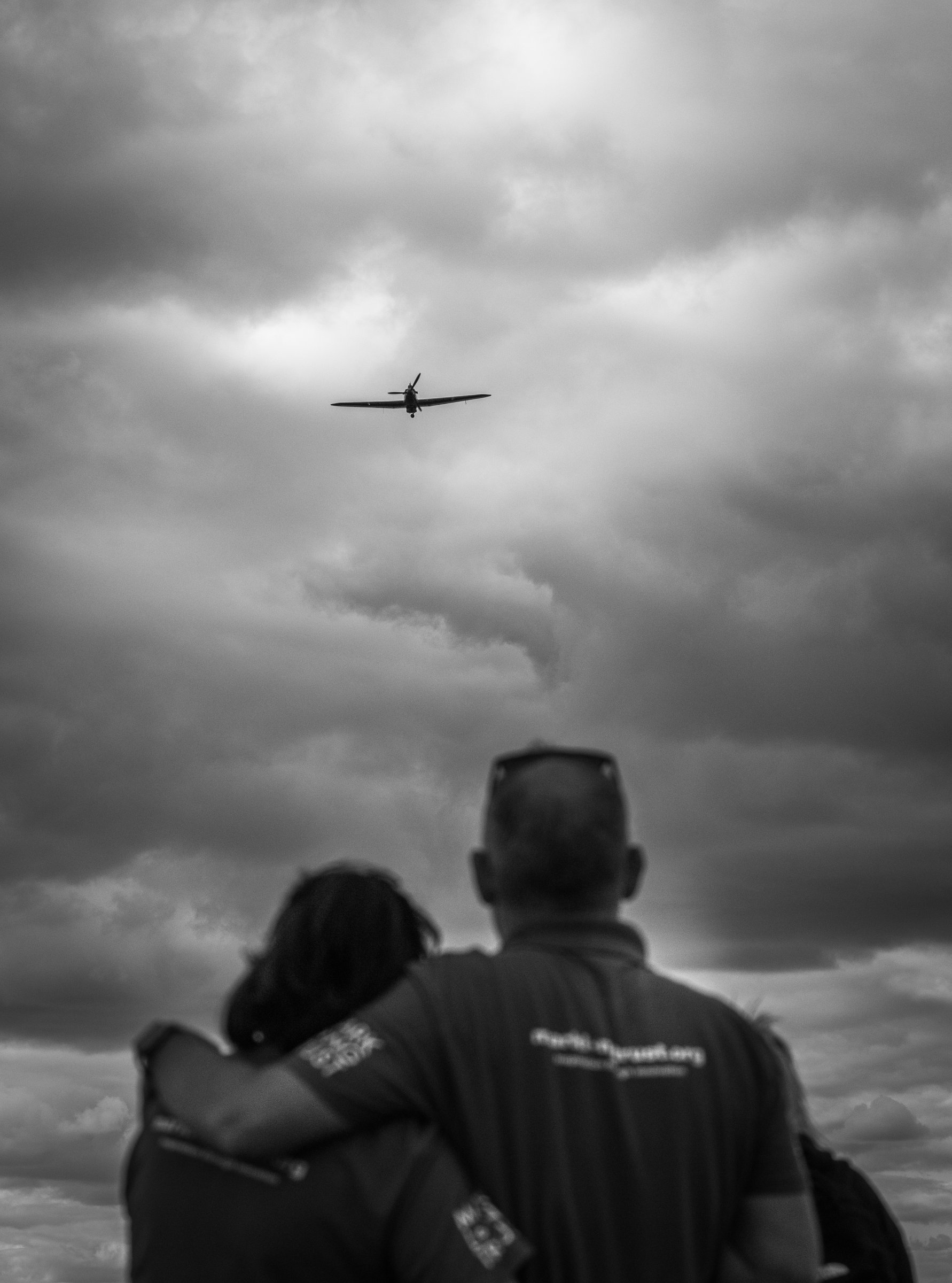 People in Mark Long Trust shirts watch a light aircraft against a cloudy sky. Credit: Peregrine Images.